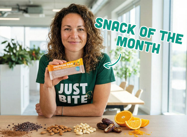 Vrouw houdt een JustBite protein bar vast aan tafel met zichtbare ingrediënten zoals amandelen, cashews, dadels en sinaasappel. Een moment van smaakvol, un-guilty snacken waarbij beter snacken moeiteloos voelt.