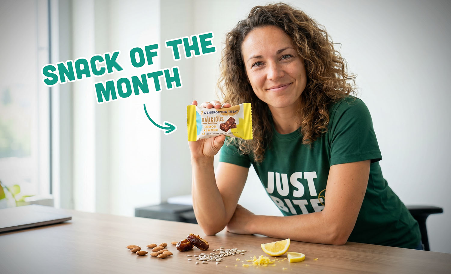 Dalicious Lemon Almond dadel-notenreep, uitgelicht als 'Snack of the Month' door een JustBite medewerker, omringd door ingrediënten zoals citroen, amandelen en zonnebloempitten.