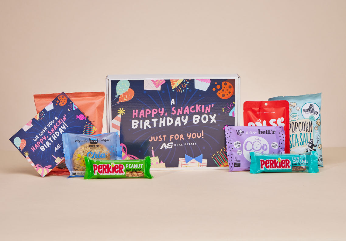Een gepersonaliseerde 'A Happy, Snackin' Birthday Box' van JustBite voor AG Real Estate, omringd door een bijpassend verjaardagskaartje en diverse bewuste snacks.
