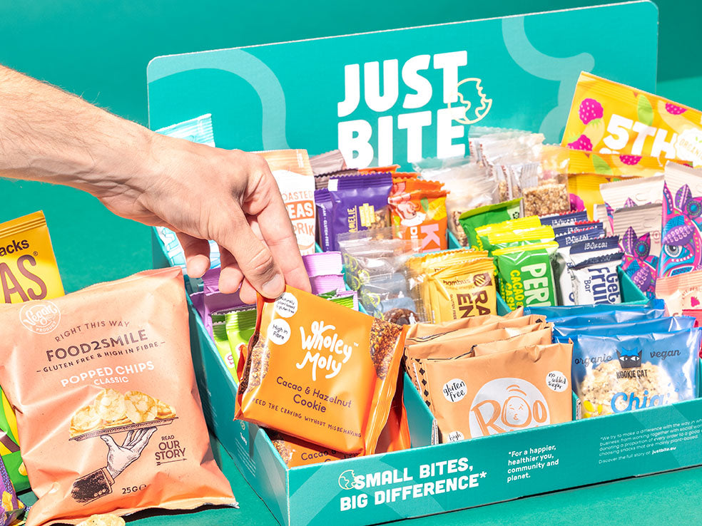 Een hand pakt een Wholey Moly koekje uit een JustBite displaydoos gevuld met een divers assortiment aan bewuste snacks.