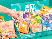 Een hand pakt een Wholey Moly koekje uit een JustBite displaydoos gevuld met een divers assortiment aan bewuste snacks.