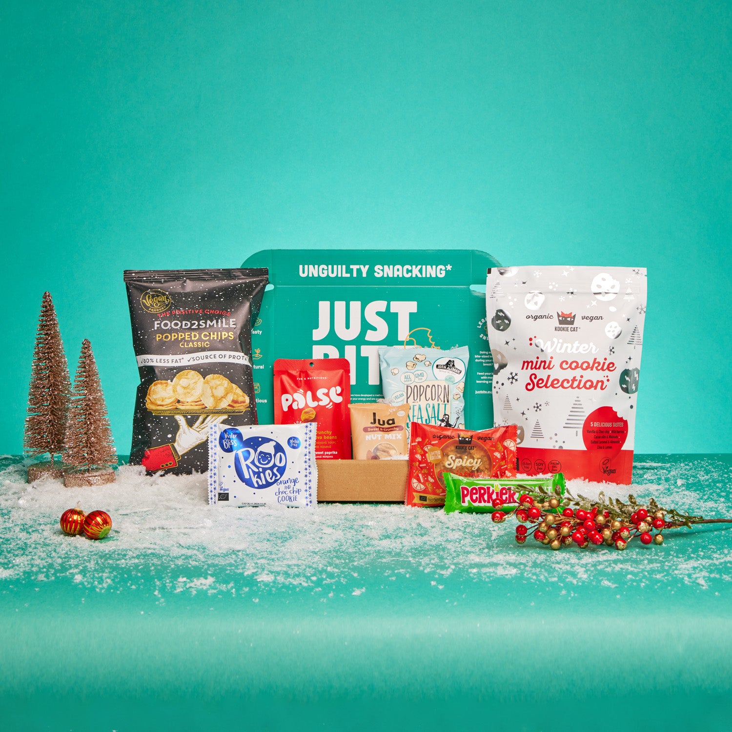 Een geopend JustBite eindejaarsgeschenk gevuld met diverse bewuste snacks, omringd door kunstsneeuw, mini-kerstboompjes en rode bessen tegen een groene achtergrond.