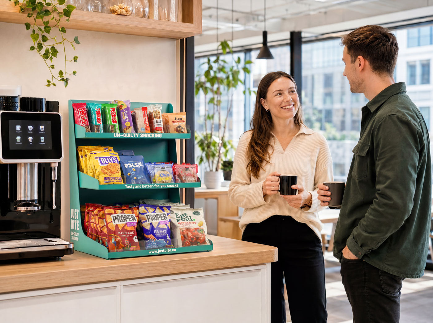 Twee collega's met koffie in gesprek naast een JustBite display met gezonde snacks op kantoor.