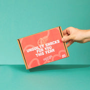 Een hand houdt een feestelijk gesloten JustBite sinterklaaspakket voor personeel vast. Op de rode doos prijkt de belofte 'Unguilty snacks for you this year'. Wij maken beter snacken makkelijk met een verrassend lekker geschenk dat goed smaakt én goed voelt.
