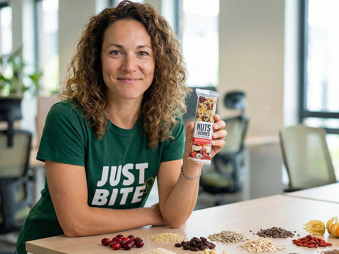 Lachende JustBite-collega toont een verrassend lekkere Nuts & Berries reep, met pure ingrediënten zoals bessen, zaden en havervlokken overzichtelijk op tafel. Wij maken beter snacken makkelijk door te focussen op 100% biologische en palmolievrije keuzes, zodat je un-guilty kunt genieten.