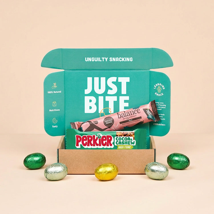 Een geopend JustBite brievenbus-paaspakket gevuld met twee bewuste snackrepen en omringd door gekleurde paaseitjes op een beige achtergrond.