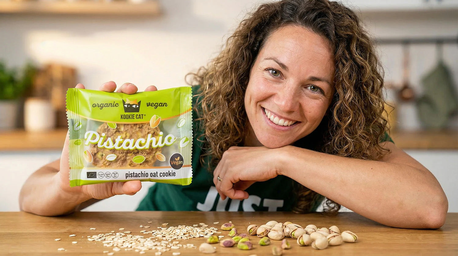 Lachende JustBite-collega toont een verrassend lekkere Kookie Cat haverkoek, met de pure ingrediënten zoals verse pistachenoten en havervlokken op tafel. Beter snacken betekent bij ons genieten van 100% clean ingrediënten, zonder verborgen troep.