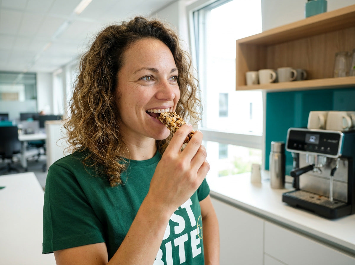 Lachende vrouw in groen JustBite-shirt eet een gezonde notenreep tijdens een pauze in de kantoorkeuken
