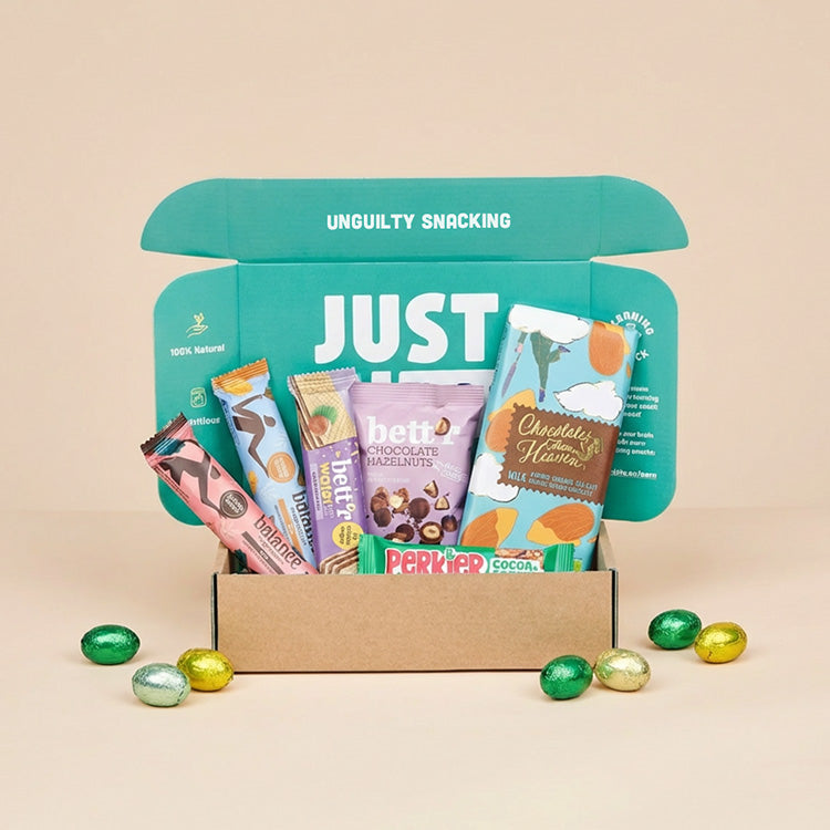 Een geopend JustBite Paaspakket Large gevuld met diverse bewuste snacks, zoals repen en chocolade, omringd door kleurrijke paaseitjes op een beige achtergrond.