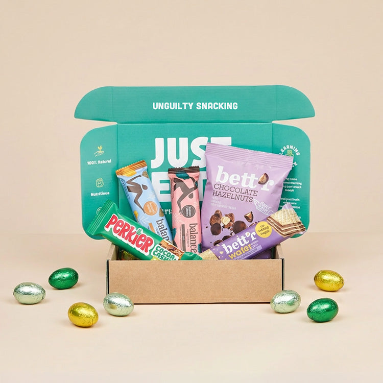 Een geopend JustBite Paaspakket Medium gevuld met diverse bewuste snacks, waaronder repen en chocolade-hazelnoten, omringd door gekleurde paaseitjes op een beige achtergrond.