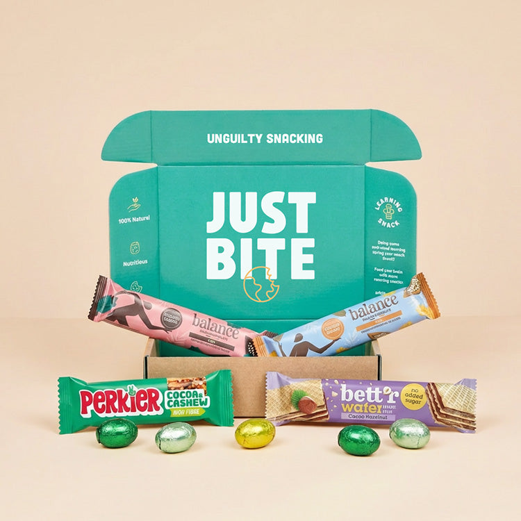 Een geopend JustBite Paaspakket Small gevuld met vier bewuste snackrepen en omringd door gekleurde paaseitjes op een beige achtergrond.