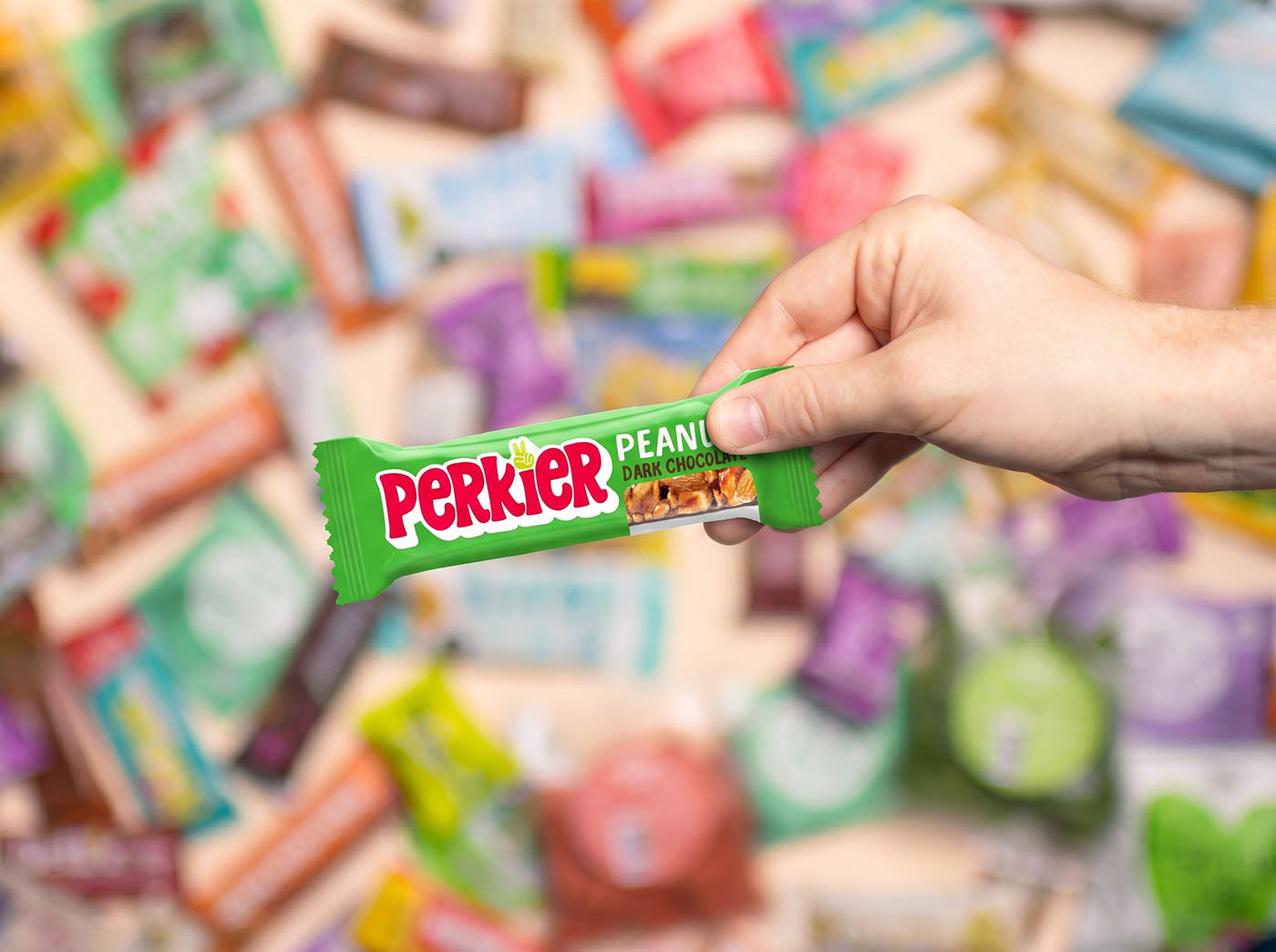 Hand houdt een Perkier Peanut Dark Chocolate reep vast, omringd door een kleurrijke selectie gezonde snacks op de achtergrond